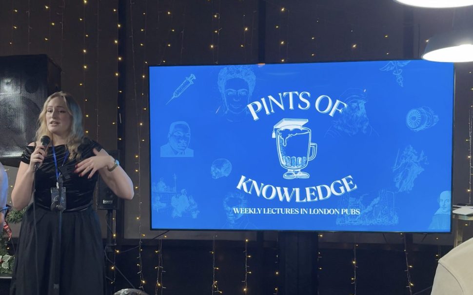 The photo depicts the logo of Pints of Knowledge with a female speaker at a microphone addressing the audience.