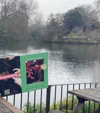 A photo of the pond in Battersea Park overlooked by the Pear Tree Cafe with some of their wooden benches and the Antiswipe sign