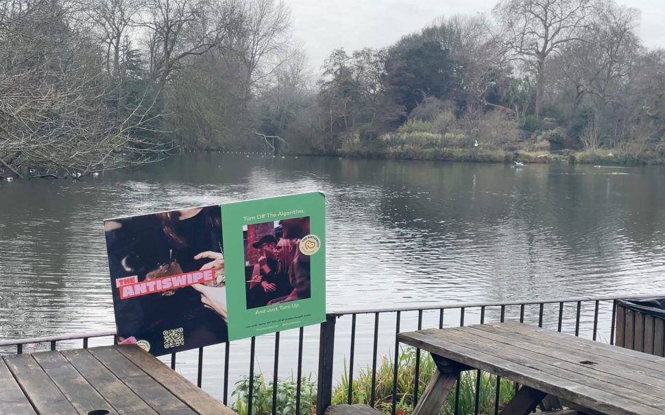 A photo of the pond in Battersea Park overlooked by the Pear Tree Cafe with some of their wooden benches and the Antiswipe sign