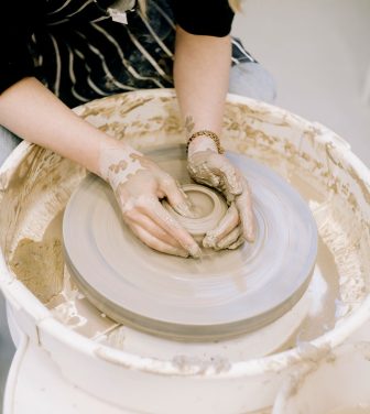 A person sitting down moulds clay on a pottery wheel with their hands