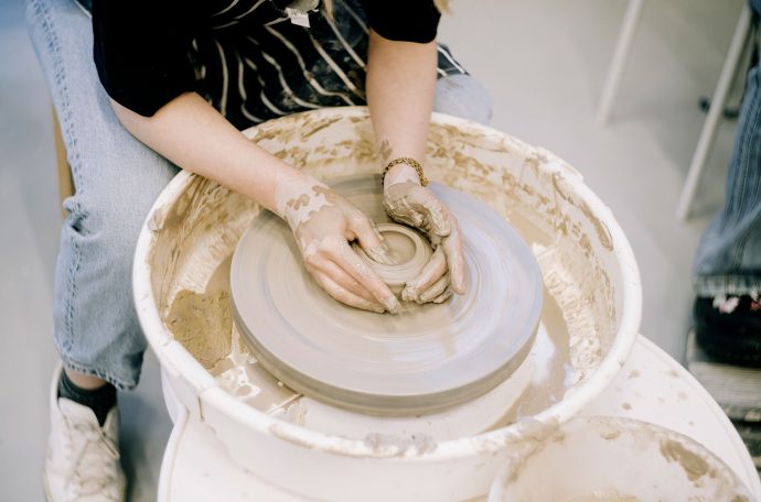 A person sitting down moulds clay on a pottery wheel with their hands