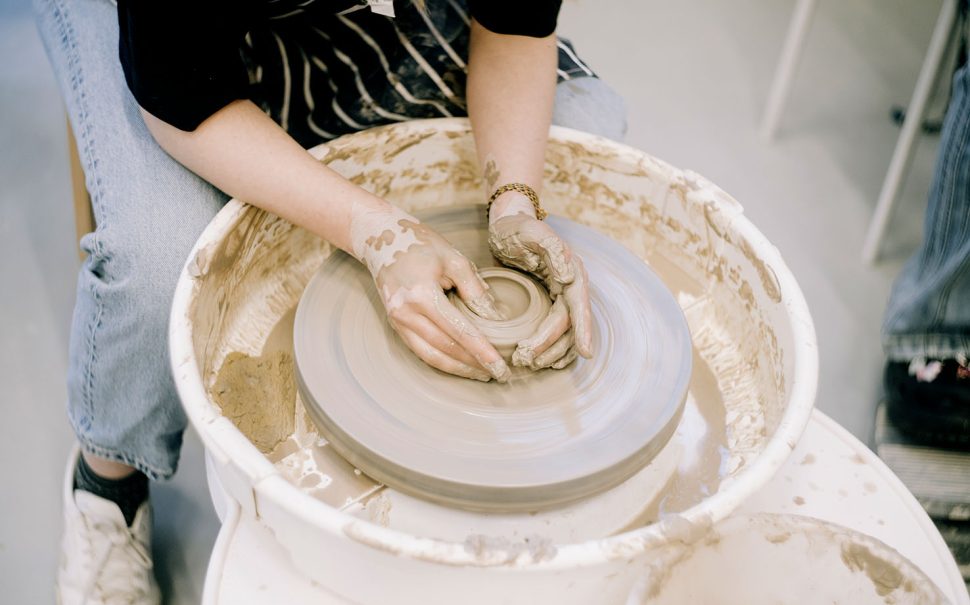 A person sitting down moulds clay on a pottery wheel with their hands