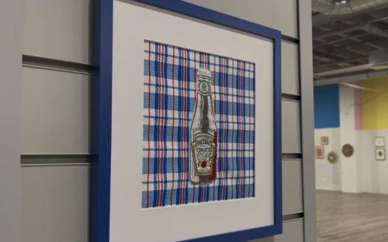 A photo showing a frame with a stitched ketchup bottle on it inside an art gallery