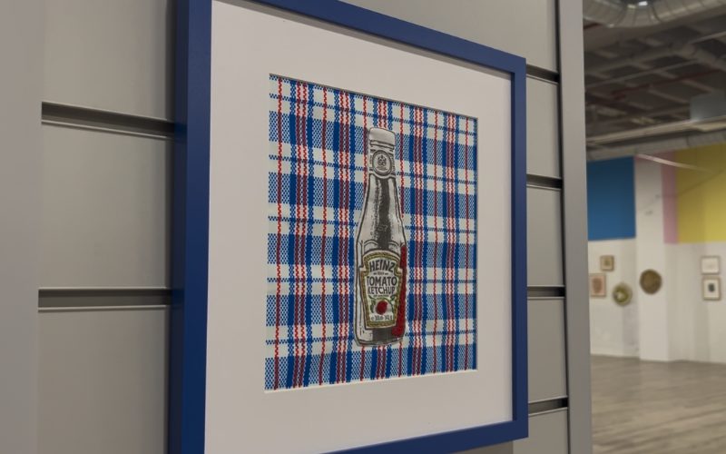 A photo showing a frame with a stitched ketchup bottle on it inside an art gallery