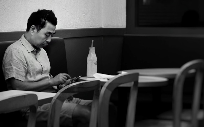 The black and white image shows a man sitting in a restaurant on his own. He sits opposite two empty chairs.
