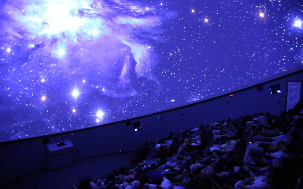 A crowd sat in cinema style seating below a planetarium projection dome.