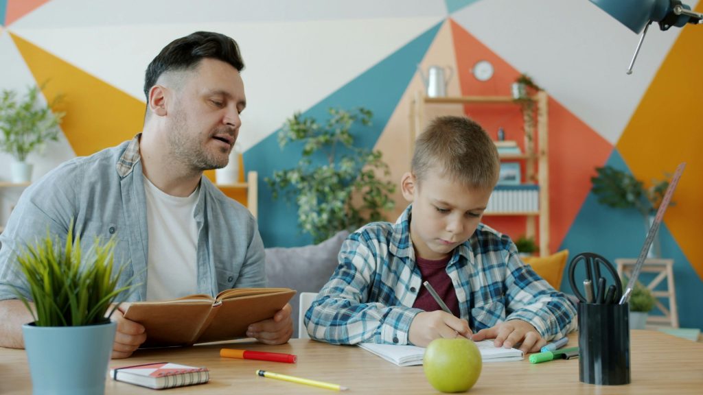 A child is doing homework with dad who is reading textbook sitting at desk at home concentrated on education.