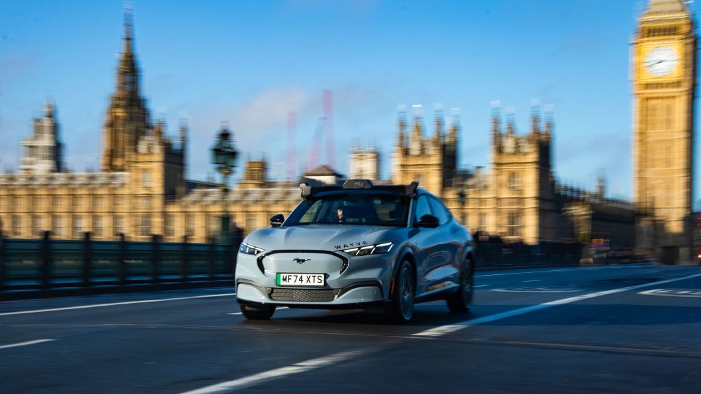 A Wayve vehicle passes the Houses of Parliament, a safety driver can be seen behind the wheel. 