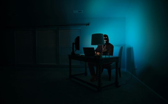 An anonymous person sitting at a computer in a dark room.