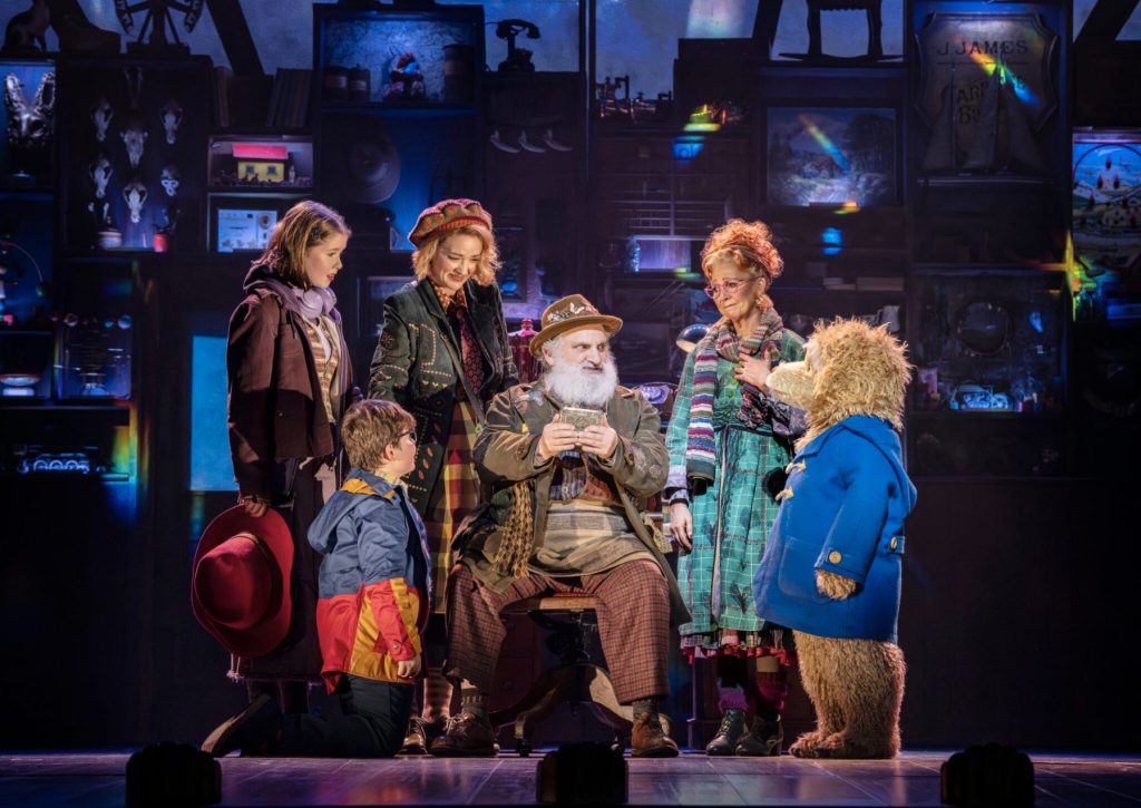 Paddington the Musical stage production with an old man sat with a young boy, three women, and Paddington bear in a blue duffel coat around him.