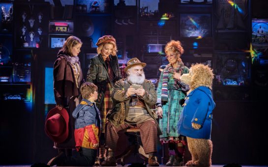 An old man sat with a young boy, three women, and Paddington bear in a blue duffel coat stood around him on stage.
