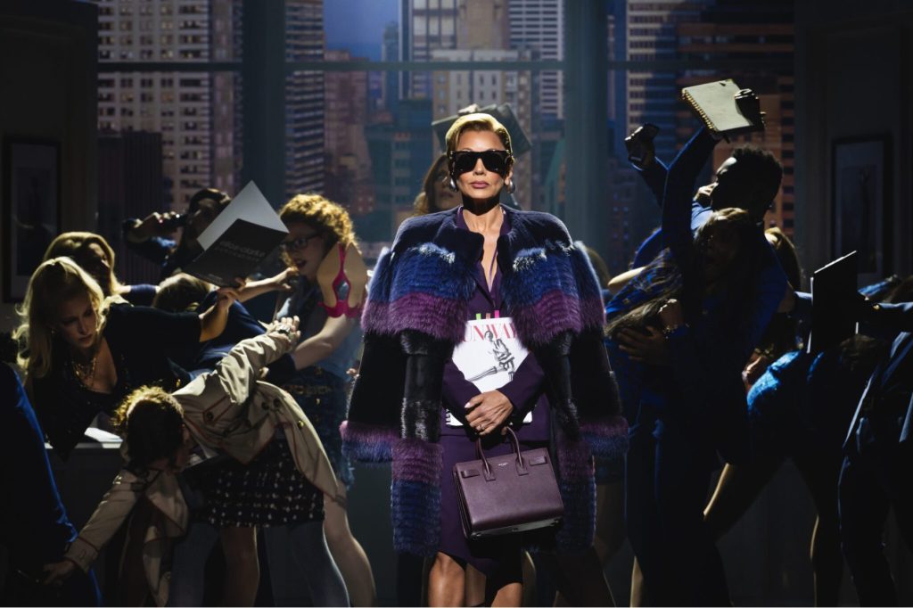 An image from The Devil Wears Prada musical of a woman wearing a fur coat and sunglasses with cast members behind her.