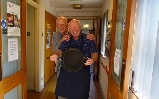 Two men, who are part of the cooking club, smile at the camera, one holding a pan.