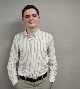 A young man in a white shirt with stripes on and dark hair stands in front of a grey background. He has his hands in his trouser pockets and he is smiling.