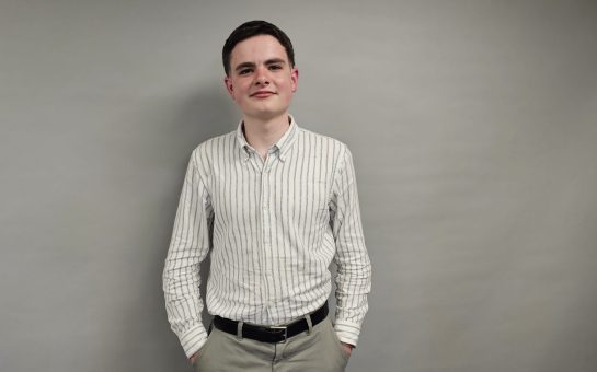 A young man in a white shirt with stripes on and dark hair stands in front of a grey background. He has his hands in his trouser pockets and he is smiling.