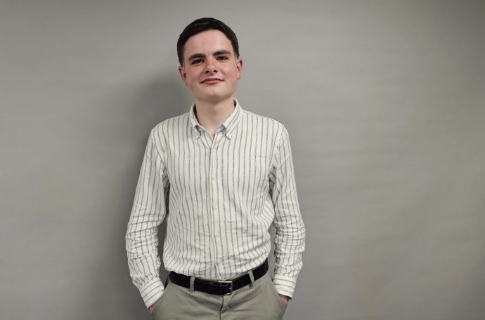 A young man in a white shirt with stripes on and dark hair stands in front of a grey background. He has his hands in his trouser pockets and he is smiling.