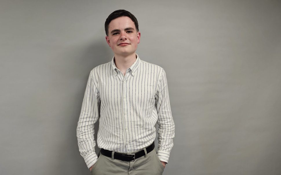 A young man in a white shirt with stripes on and dark hair stands in front of a grey background. He has his hands in his trouser pockets and he is smiling.