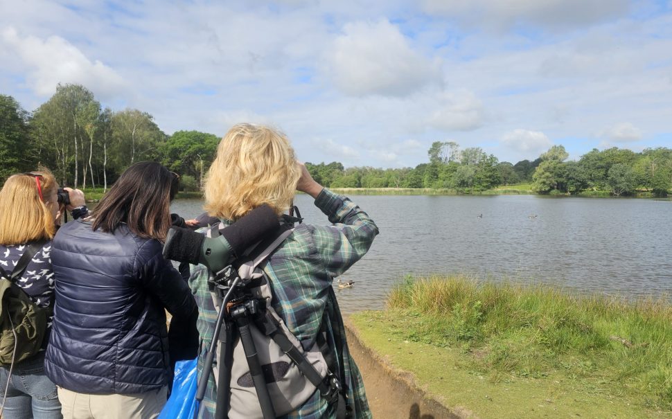 3 women with binoculars looking out over a lake in Richmond Park. They are bird watching.