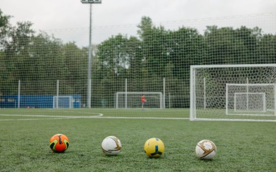 Four footballs lined up on a pitch with two goals in the distance.