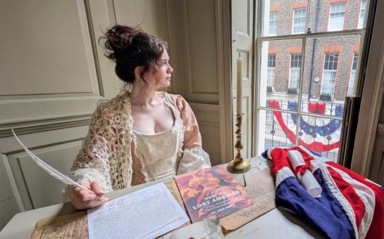 A stylised portrayal of the Declaration of Independence being signed. A woman sits with a quill looking out of the window, with an early US flag on the table and draped from the balcony.