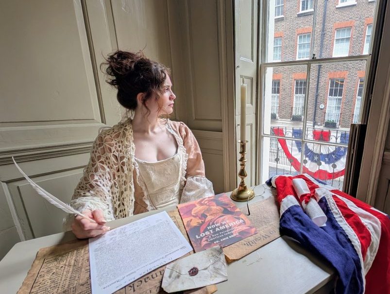 A stylised portrayal of the Declaration of Independence being signed. A woman sits with a quill looking out of the window, with an early US flag on the table and draped from the balcony.