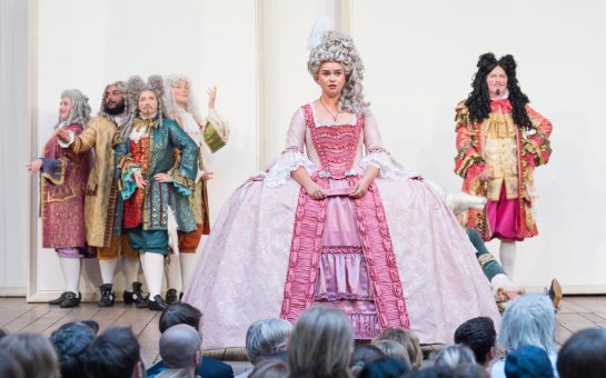 A leading actress stands at the front of the stage at Shakespeare's Globe in a regency era gown.