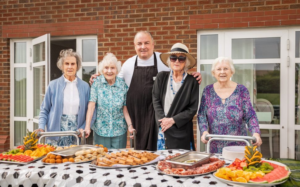 Residents of a care home stand beside some food on a table, with a chef in the middle.