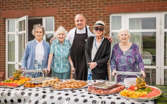 Residents of a care home stand beside some food on a table, with a chef in the middle.