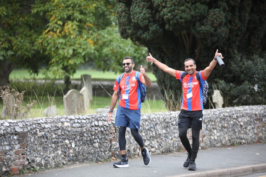Two Marathon March walkers in red and blue tops walk on road with arms raised, thumbs up and smiles on faces.