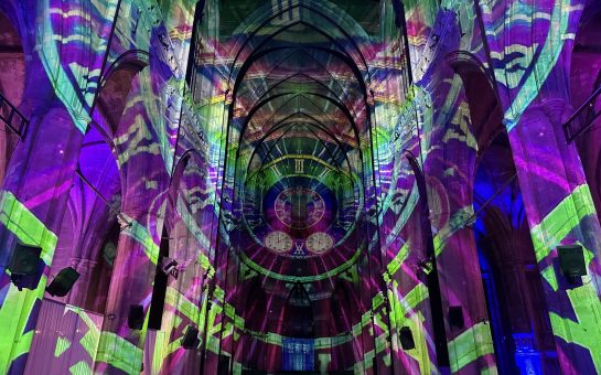 The building walls are covered in projections of clock faces layered over one another. They are rainbow coloured and all lead into one clock that acts as a focal point in the middle of some grand arches.