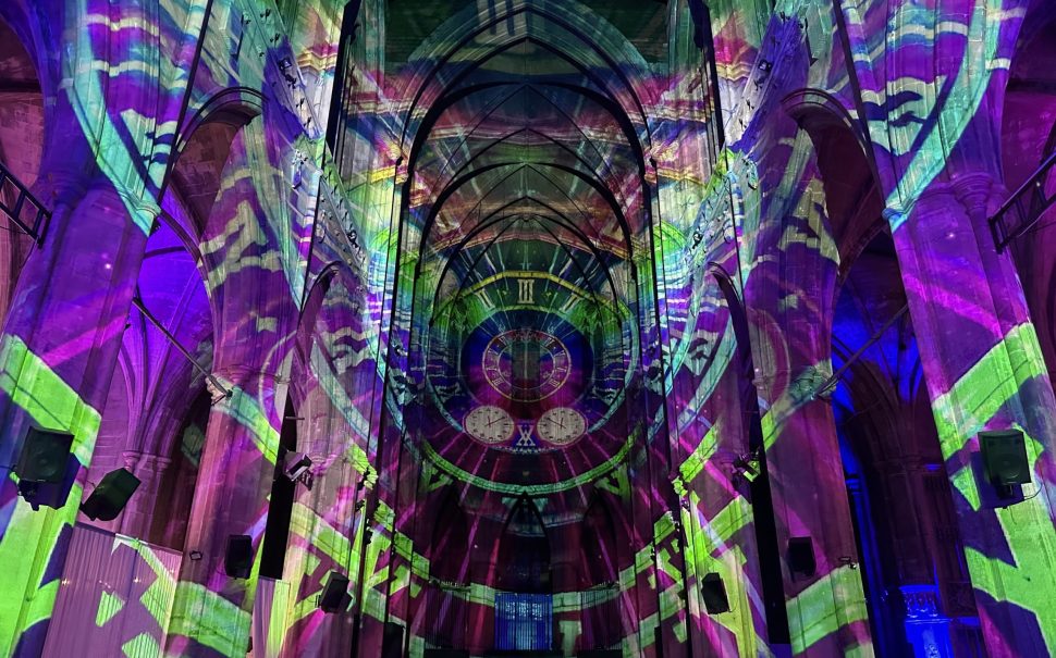 The building walls are covered in projections of clock faces layered over one another. They are rainbow coloured and all lead into one clock that acts as a focal point in the middle of some grand arches.