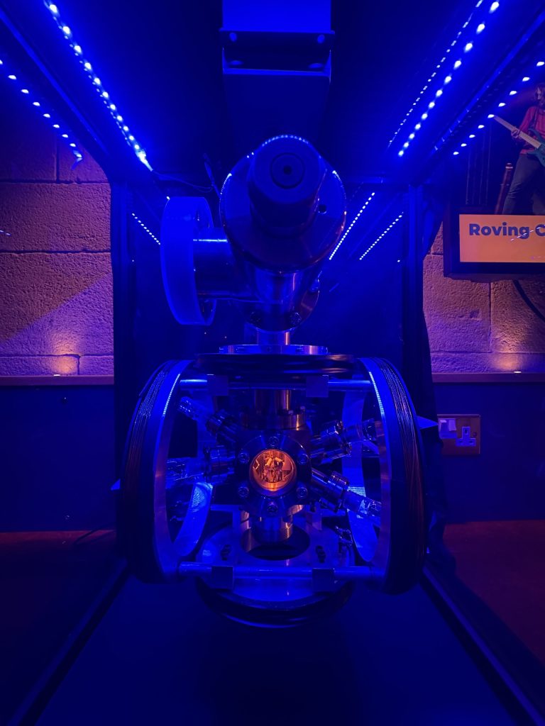A close up shot of part of an atomic clock. It appears to be made of metal structures and cogs. It has a small hole in the centre with a lots of little wires, lit up in orange.