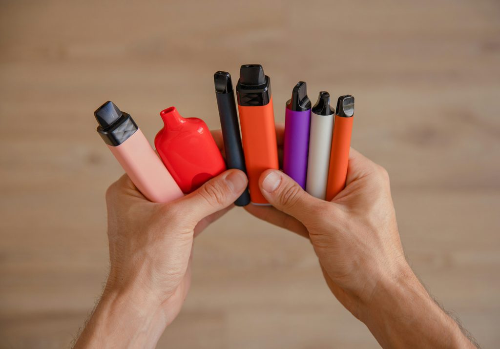 A pair of hands holding up seven vapes, all different sizes and colours.