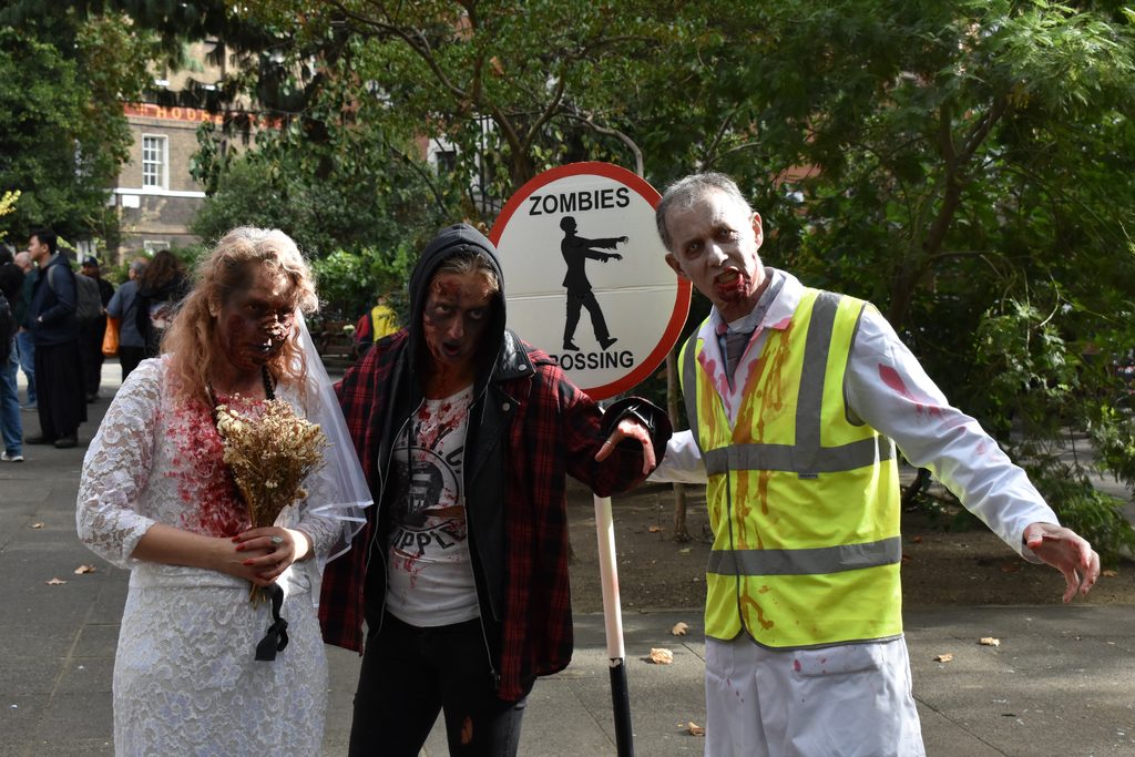 Steward, dressed as a zombie, holds a sign saying 'Zombies Crossing' next to two people dressed as zombies.