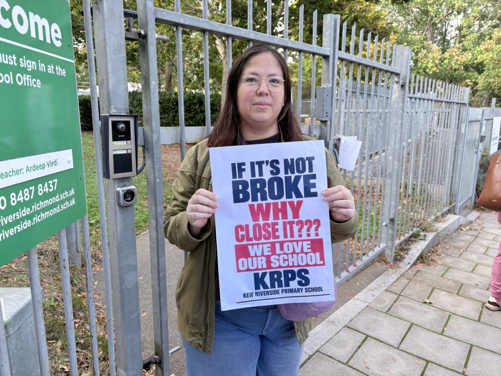 photo of Cecilia Webb standing in front of kew riverside primary school holding a sign saying "If it's not broke why close it?"