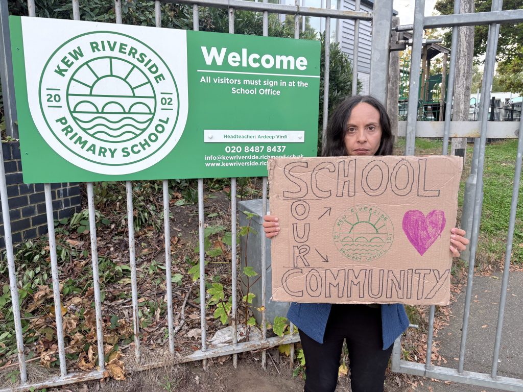 a photo of Priscila Smith holding a sign outside kew riverside primary school that says "School - Our Community"