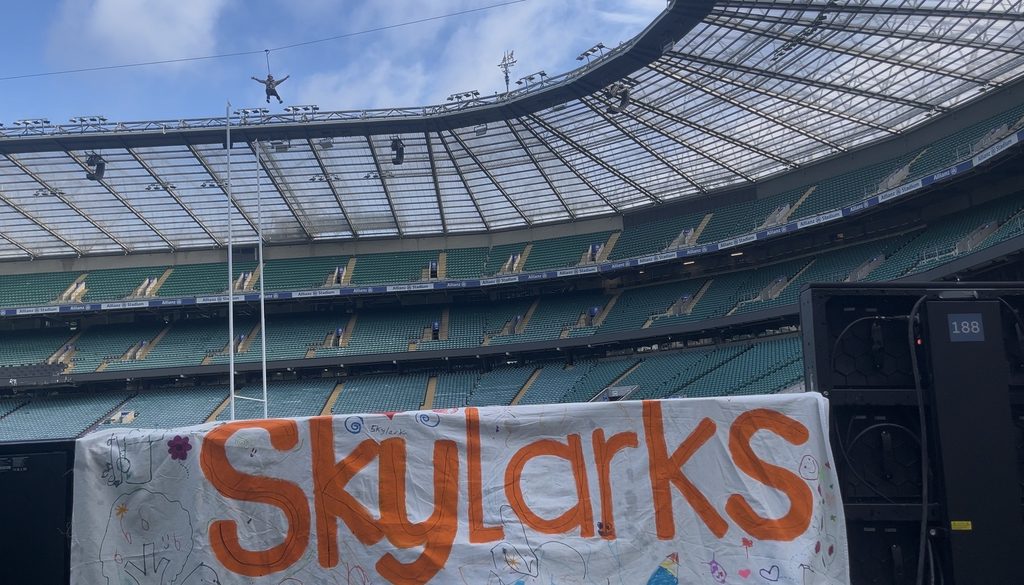 A zipliner flies over the rugby pitch at the Allianz stadium with a Skylarks charity banner below.