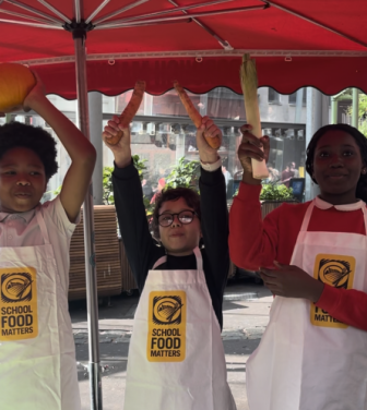 Three children raise vegetables above their heads at a market.