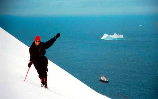 An image of Catherine scaling a mountain in Antartica, holding climbing gear and wearing winter clothes as she smiles and waves for the camera