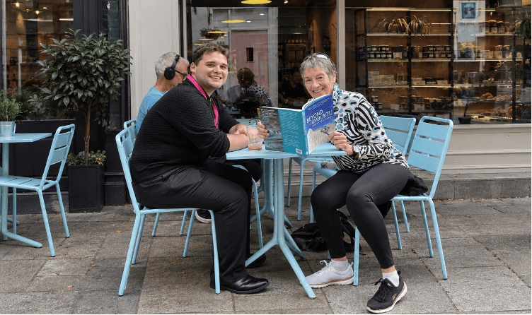 A photo of Catherine DeVrye sat at a coffee shop holding open her book, reading it with a journalist.